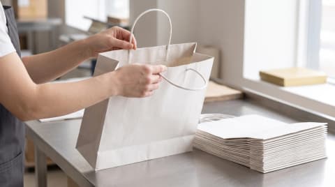 Hands checking the handle of a white paper bag beside a stack of flat bags